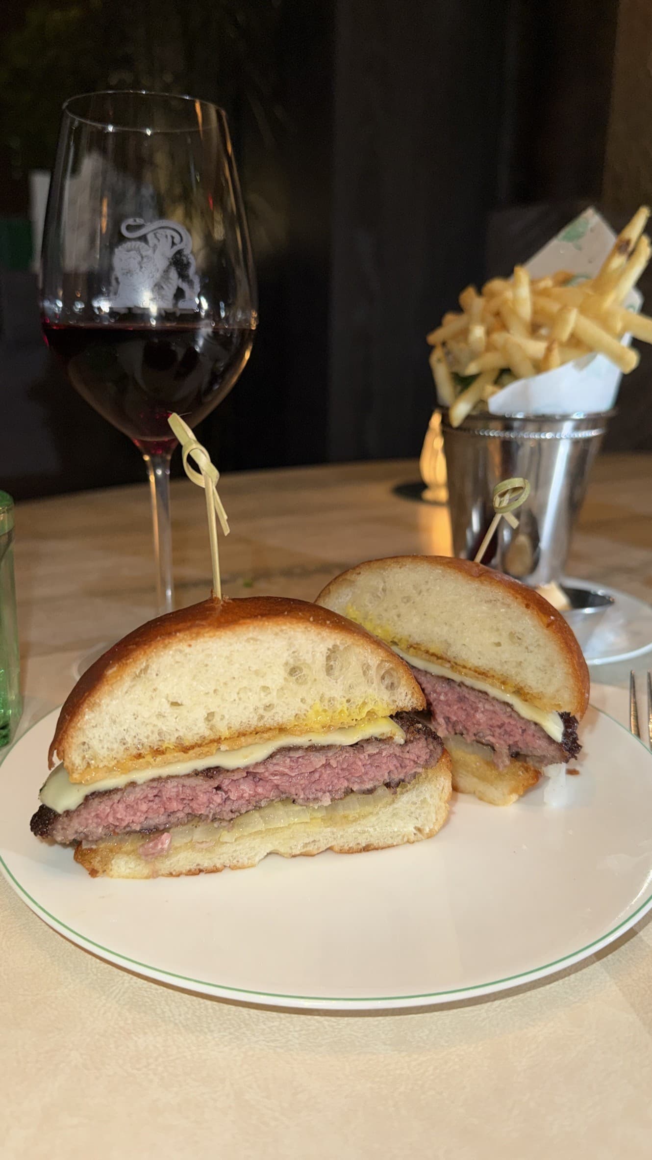 The 550 Burger at Bar Chimera, halved to show a rosy interior, with melted cheese, a Parmesan crisp and mustard under a domed brioche bun, beside tallow fries in a silver cup