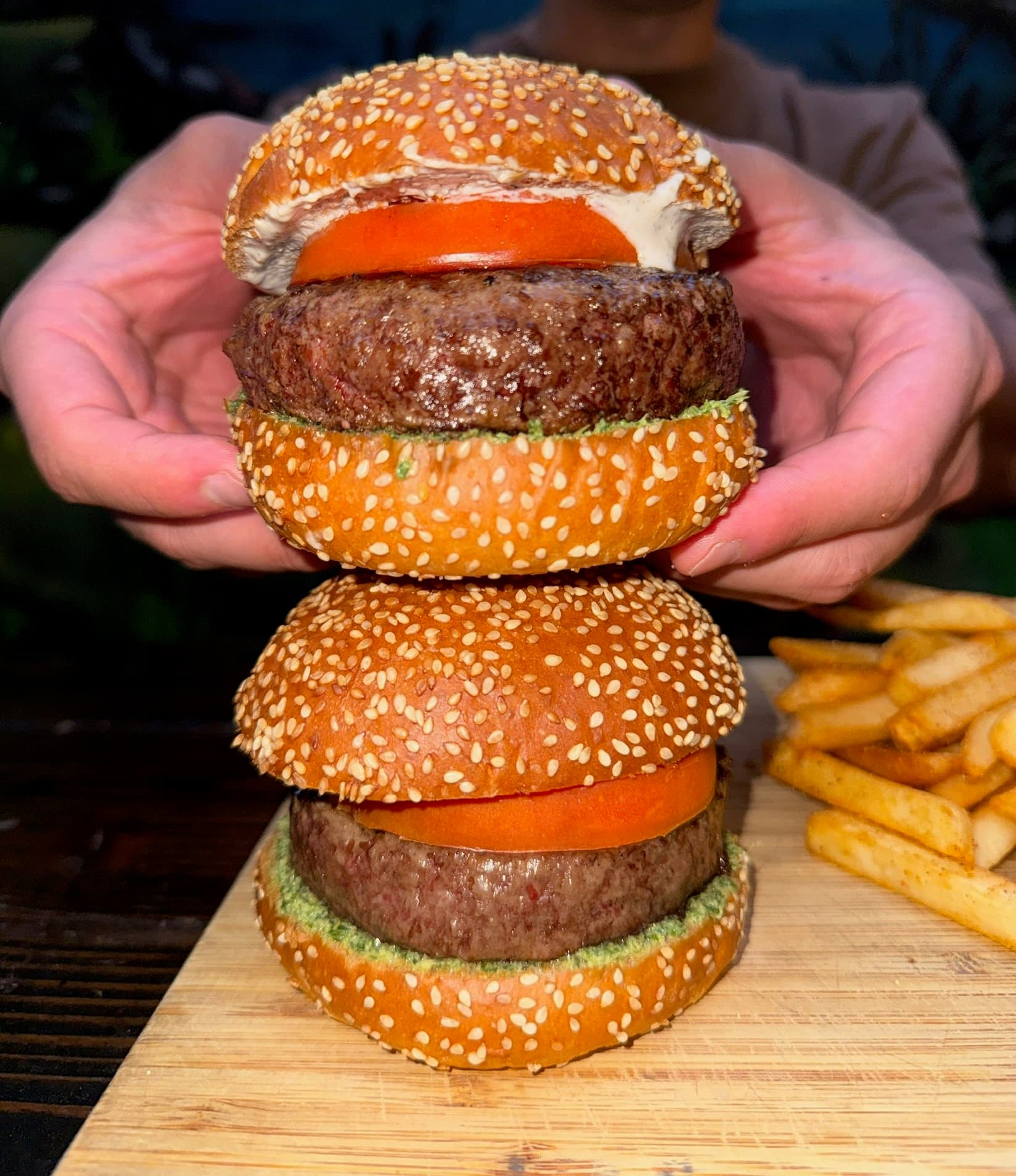 Two of BK Jani's Jani burgers stacked in a pair of hands: thick patties over green mint chutney, with a slice of tomato and raita on sesame buns, and fries on a wooden board