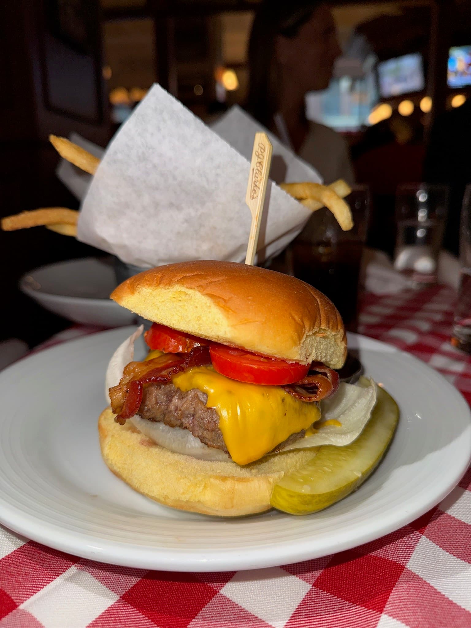 The Cadillac at P.J. Clarke's Manhattan West on a red-and-white checked tablecloth: a patty with melted American cheese, bacon, tomato and lettuce on a potato roll, held with a P.J. Clarke's pick, a pickle spear on the plate and fries behind