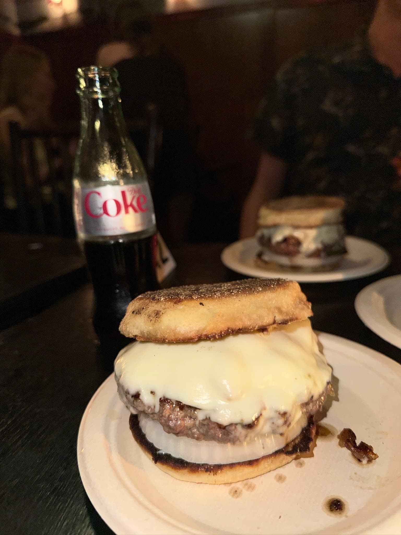 The Shy's Burgers and Stone & Skillet English-muffin burger at The River, on a paper plate: a thick patty under melted white cheese over a slice of raw onion, with a Diet Coke bottle and a second burger behind