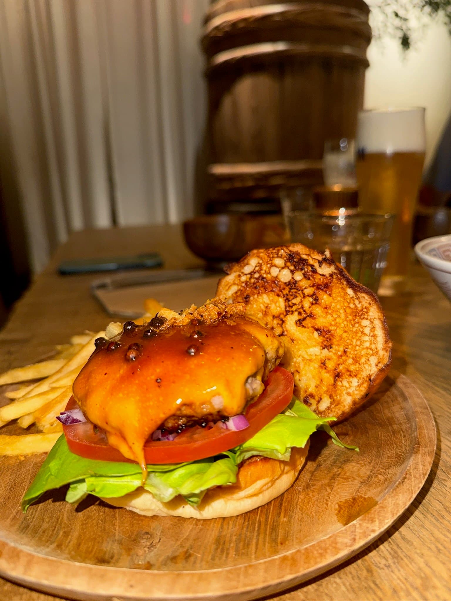 THE GALLERY by Odo's burger served open on a wooden plate: a patty under melted orange cheese glazed with sauce and whole peppercorns, crispy fried onion, tomato, lettuce and diced red onion, the charred top bun beside it with fries and a beer