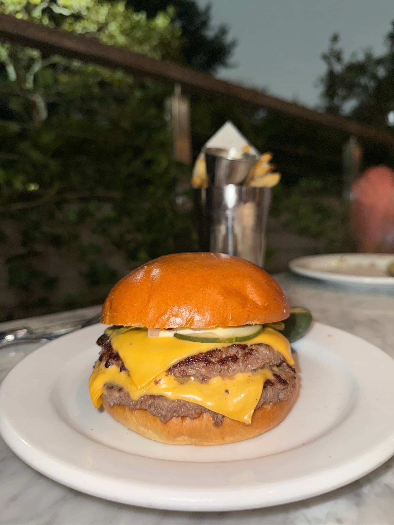 The Snail's Double Cheeseburger at an outdoor table at dusk: two patties with melted American cheese and pickle slices under a glossy brioche bun, with fries in a steel cup behind